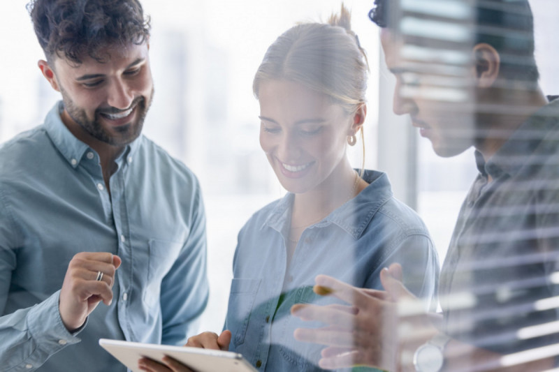 Business-Team schaut lächelnd gemeinsam auf ein Tablet – legere Atmosphäre, Fenster im Hintergrund